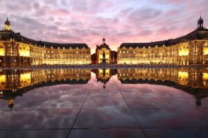 Vue panoramique de la Place de la Bourse à Bordeaux avec le Miroir d'eau reflétant les façades illuminées au crépuscule